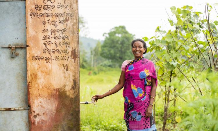 Raibari Bewa standing near the toilet, bathroom unit and collecting water from the third tap in Dudukaguda village, in Thuamul Rampur block, Kalahandi district of Odisha. On the walls, details of Swachh Bharat Mission benefits availed by her in Odia | Picture courtesy: Ajaya Behera